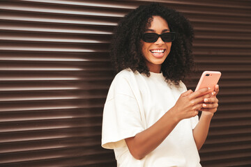 Beautiful black woman with afro curls hairstyle.Smiling hipster model in white t-shirt. Sexy carefree female posing in the street near brown wall in sunglasses.Looking at smartphone screen, using apps
