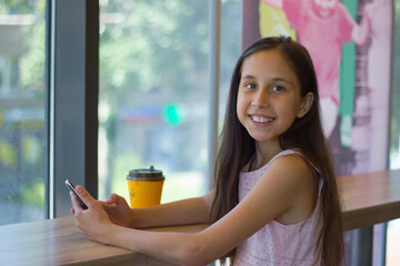 A girl with long dark hair sits in a cafe, drinks tea, holds a mobile phone and looks at the camera. 