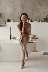 Joyful brunette Asian lady smiles sincerely, leans on her knees, looks into camera, holds magazine...