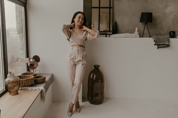 Tanned brunette Asian woman in linen pants and cropped beige top leans on wall in cozy bathroom and smiles sincerely.