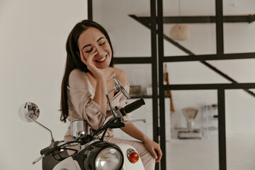 Obraz na płótnie Canvas Pretty brunette Asian woman in stylish beige linen blouse and pants smiles widely, touches her face and sits on moped in apartment.