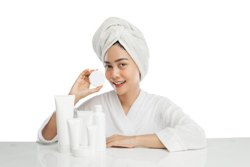Smiling beautiful girl model with towel on head holding cream pot bottle with various skin care products on table on light gray background