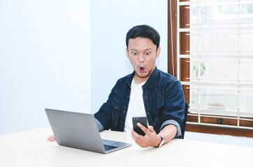 Fototapeta premium Young Asian man shocked and wow what he see in the laptop when in call on smartphone
