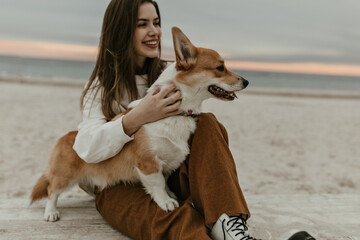 Charming brunette woman in brown pants smiles sincerely, sits on beach and plays with corgi in good...