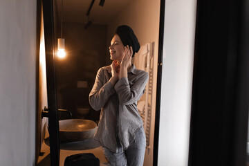 Attractive lady in black towel on head looks into mirror in bathroom. Beautiful woman in grey shirt and jeans does skin care routine.