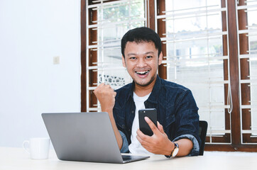 Fototapeta premium Happy excited and smiling young Asian man raising his arm up to celebrate success or achievement when working in laptop. Indonesian man wearing blue shirt.