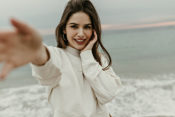 Pretty elegant woman in white sweatshirt smiles sincerely, gently touches her face and takes selfie on sea background.