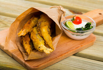Fried sprats in batter served on wooden cutting board with cheese sauce, lime and tomato