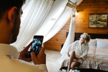 The groom takes a picture on the phone: woman wearing a garter on the leg. The bride holds a hand garter and hotel room. morning preparation wedding concept.