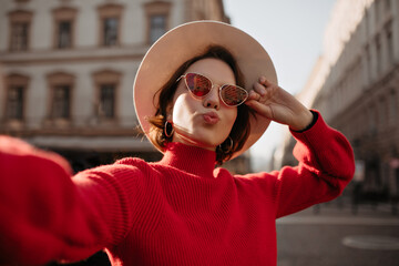 Short-haired woman in red sweater and sunglasses takes selfie outside. Beautiful lady in beige hat...