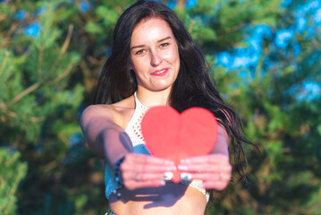 Pretty brunette showing red heart smiling dreamy nature summer sunny day.Woman in nature holds red paper heart in front