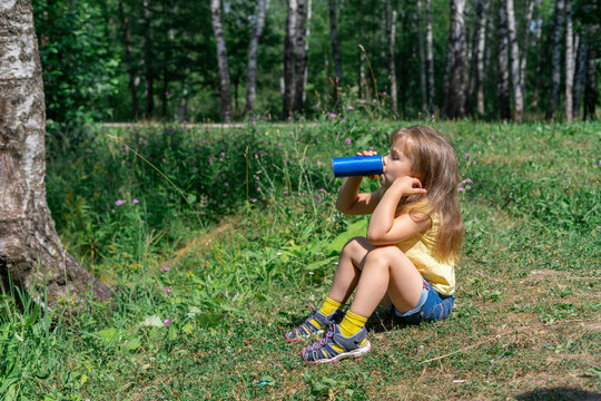 little girl drinking soda from a metal can by the lake while sitting on the grass on a hot summer day