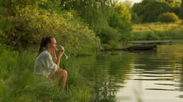 Young woman enjoying glass of wine on lake bank at sunset. Woman admiring landscape while having drink