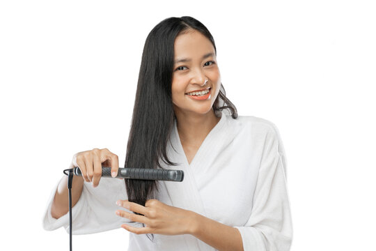 Beautiful girl in white towel using a hair straightener and smiling while looking the camera in gray background