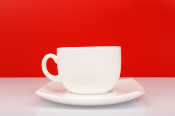 Close up of white ceramic coffee or tea cup with saucer on white table against red background. Concept of hot drinks and kitchenware