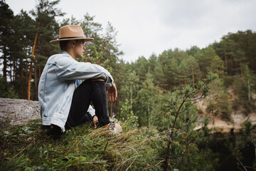 Side view low angle of young male traveler sitting on stony edge and admiring scenery of lake surrounded by coniferous forest