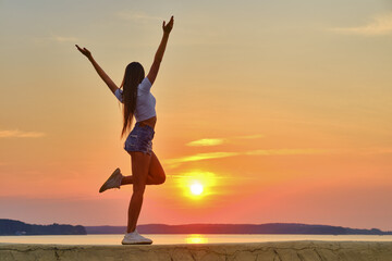 Young woman jumping and spreading hands to the sides and facing to the sunset