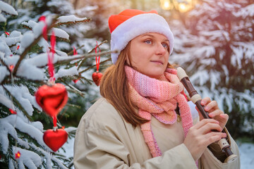 A happy woman in winter clothes plays the flute at the Christmas tree in a snow park on New Year Eve