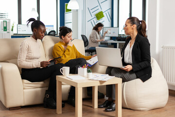 Group of multiethnic business people discussing about company strategy sitting on coach in modern cooperation office looking at statistics. African entrepreneur having a meeting with investors.