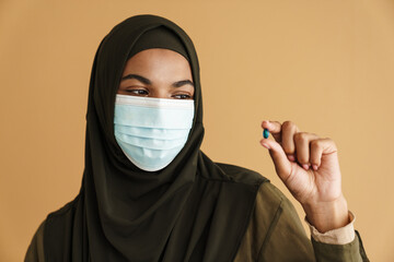 Black muslim woman in face mask showing and holding vitamin capsule