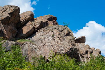 Granite rocks close-up on background blue sky with white clouds, green leaves on treetops.