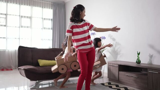 excited two sister playing airplane and car made of cardboard box running around the house with mom