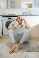 A gray-haired man sitting on the floor in the bathroom and looking upset