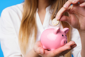 Obraz na płótnie Canvas Female putting money into pink piggy bank against blue background