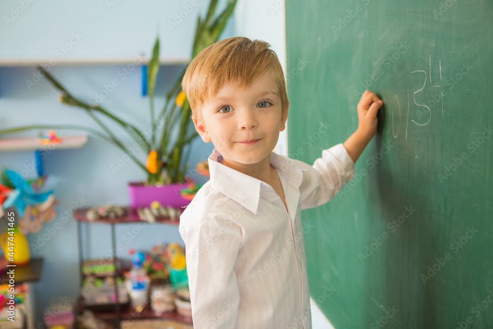 Wall mural A little cute boy at school is standing near the blackboard and writing on it. Back to school concept