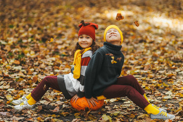 Happy playful children friends in warm clothes a hat in bright hats and scarves are walking having fun sitting on a stump and a pumpkin catching leaves in the autumn forest in nature, selective focus