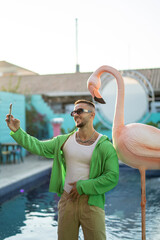 Chico joven atractivo vestido a la moda tomandose selfies frente a una piscina y un flamenco...