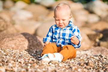 A little baby boy in a blue shirt plays with stones and shells on the beach