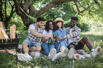 Fototapeta premium Young cheerful men and women sitting together on checkered plaid at summer garden and drinking cold beer. Multicultural friends enjoying picnic time on fresh air.