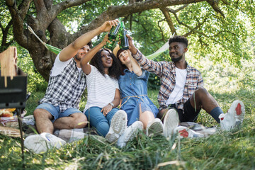 Multiracial group of friends in summer clothes drinking beer during picnic at green garden. Young happy people relaxing together on nature . Concept of friendship and enjoyment.