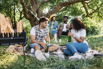 Group of four joyful friends in casual clothes having picnic at summer garden. Two multiracial couple sitting on plaid and hammock, talking and drinking alcohol.