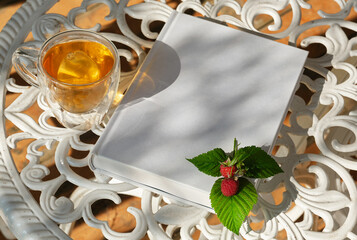Transparent glass cup of herbal tea against the background of a white garden table in the garden. White book with raspberries and red currants. Shallow depth of field