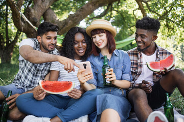 Group of young diverse friends in casual clothes sitting together at green garden and looking on smartphone screen. Four people drinking beer and eating watermelon outdoors.