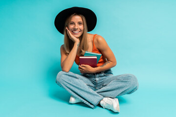 Cute pretty female student sitting on floor with books in studio over blue background.  Wearing...