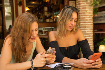 Two women sitting on the terrace of a bar checking social networks on their cell phones.