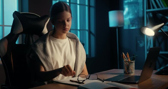 Smart teenage student studying at home late at night, feeling tired of writing, taking off eyeglasses and rubbing her itchy eyes.