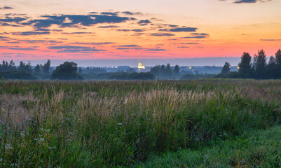 Colorful sunrise in rural with bright sky in the background colored by rising sun. A church and trees look out from a foggy meadow. Dusk and dawn, agriculture, travel, tourism, scenic views, hiking.