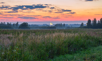 Obraz premium Colorful sunrise in rural with bright sky in the background colored by rising sun. A church and trees look out from a foggy meadow. Dusk and dawn, agriculture, travel, tourism, scenic views, hiking.