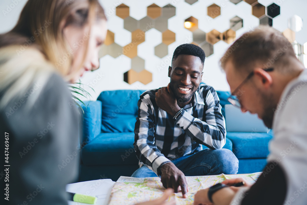 Poster smiling black man pointing at place on map to colleagues at coffee table