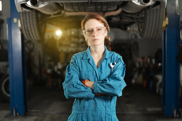A young woman in work clothes, an apprentice stands next to the car and is proud and happy in the garage. holding a wrench in his hands. - obrazy, fototapety, plakaty