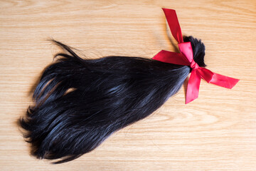 Mechón de pelo cortado anudado con un lazo de raso rojo sobre un fondo de madera. Donación de cabello para pelucas para pacientes con cáncer - obrazy, fototapety, plakaty