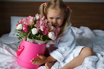 Little girl in a robe holds a box of flowers while sitting on the bed