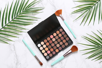 Eyeshadow palette with makeup brushes on marble background with palm leaves