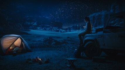 Female Traveler Sitting Uses Smartphone, Gazing at Starry Night Sky while Camping in the Canyon by...
