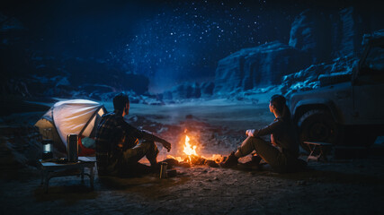 Happy Couple Sitting by Campfire Watching Night Sky while Camping in the Canyon. Two Traveling people Tell Inspirational Stories at Campsite, Look at Milky Way Stars. On Holiday Vacation Trip - obrazy, fototapety, plakaty