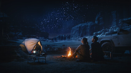 Happy Couple Tent Camping in the Canyon, Sitting by Campfire Watching Night Sky with Milky Way Full of Bright Stars. Two Travelers In Love On a Romantic Vacation Trip. Back View Shot - obrazy, fototapety, plakaty